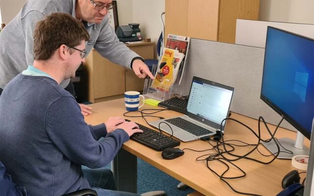 Younger man at laptop with older man helping